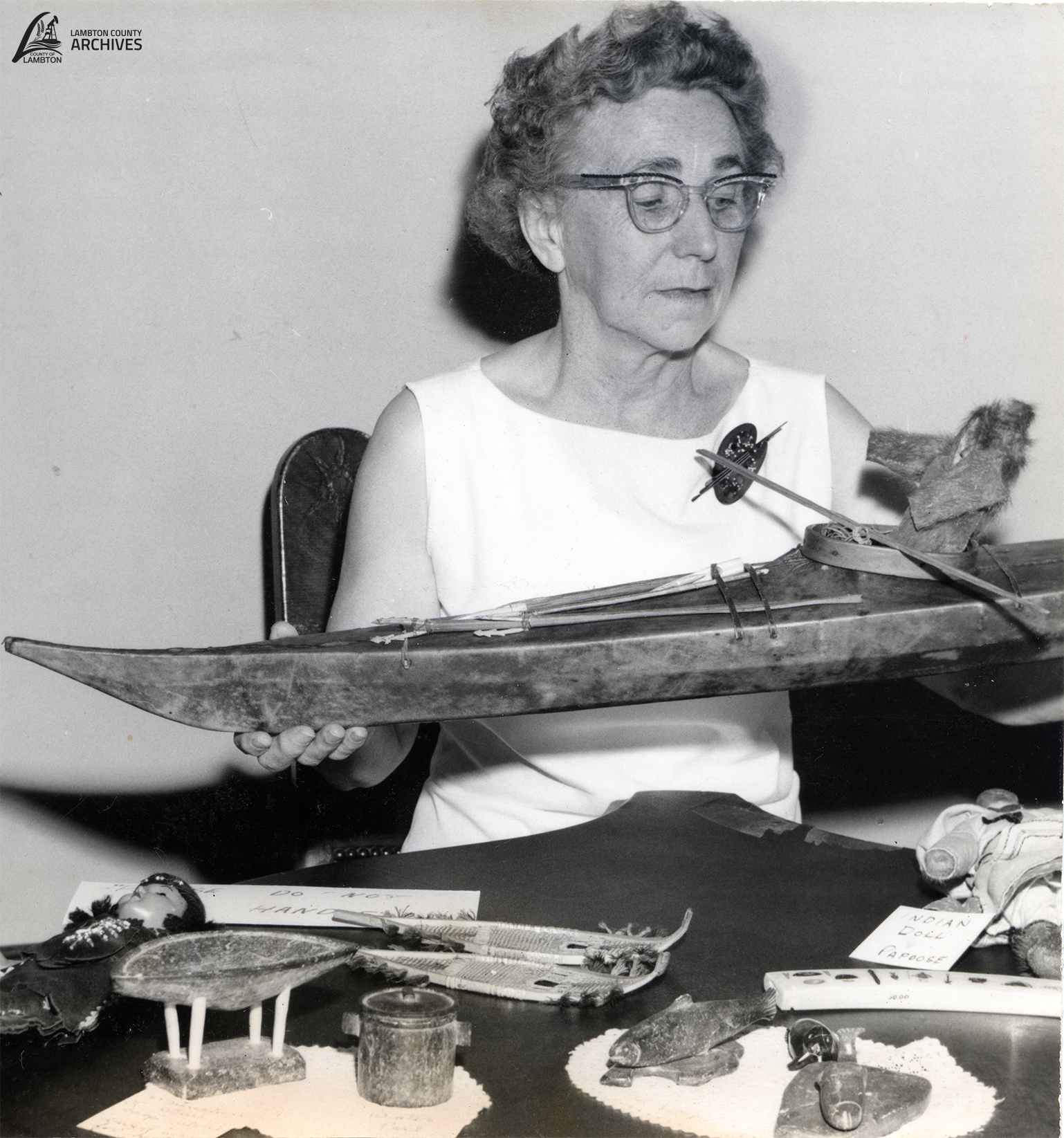 A black-and-white photo Gladys Slack, seated at a table displaying various Indigenous artifacts. She is holding a model canoe, and there are other items in front of her, including miniature snowshoes, a doll, and small containers. The photo is watermarked with "Lambton County Archives" in the top-left corner.