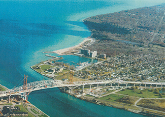 he image shows a turquoise blue waterway with urban development along the shoreline. There's a prominent bridge structure spanning across the water, connecting two land masses. The landscape includes both developed areas with buildings and some less developed or open spaces. 