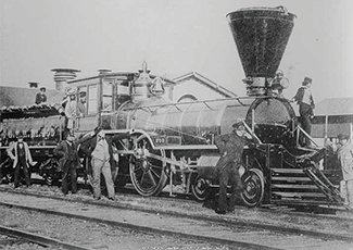 A vintage black and white photograph shows railroad workers gathered around a steam locomotive, with its distinctive funnel-shaped stack and detailed mechanical components clearly visible.