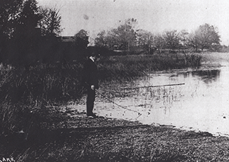 A black and white photograph captures a solitary figure fishing at the edge of a serene lake or pond, surrounded by trees and reeds in what appears to be a peaceful rural setting.  Copy Re