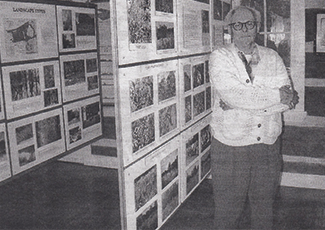 a person standing in what appears to be a gallery or exhibit space. They are dressed in a cardigan and glasses, standing with arms crossed. Behind them are display boards filled with photographs or informational materials, organized in a grid format. The setting suggests a historical or educational exhibit.