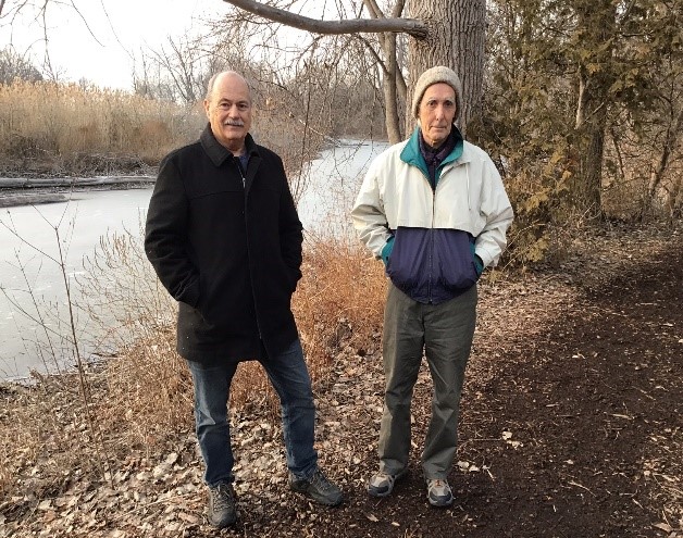 Two men are standing outdoors on a dirt path near a frozen river, surrounded by winter trees and dry vegetation. One is wearing a dark coat and jeans, while the other is in a white and blue jacket with a beanie and green pants. The setting is calm and overcast.