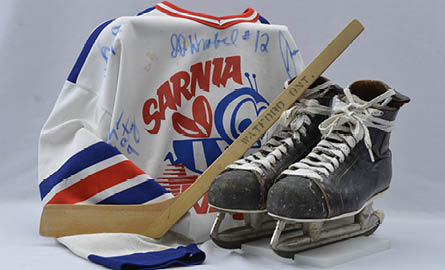 A hockey jersey, mini stick, and hockey skates grouped together with a white background.
