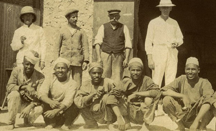 Row of Egyptian men sitting on ground with row of men in white suits standing behind.