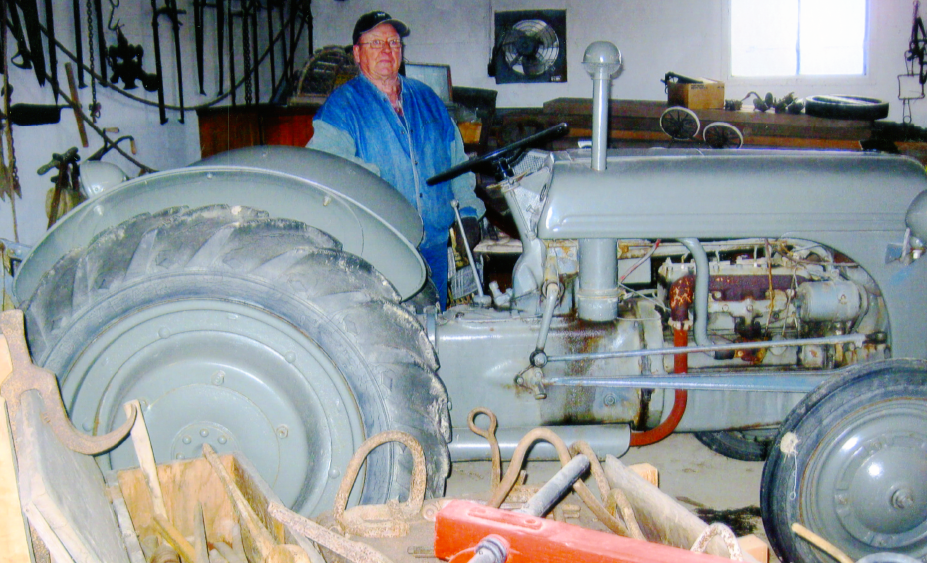 Man in blue coveralls standing next to antiques and a tractor. 