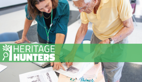 Two people pointing at photos and documents on a table, with a green translucent overlay that displays the "Heritage Hunters" logo.